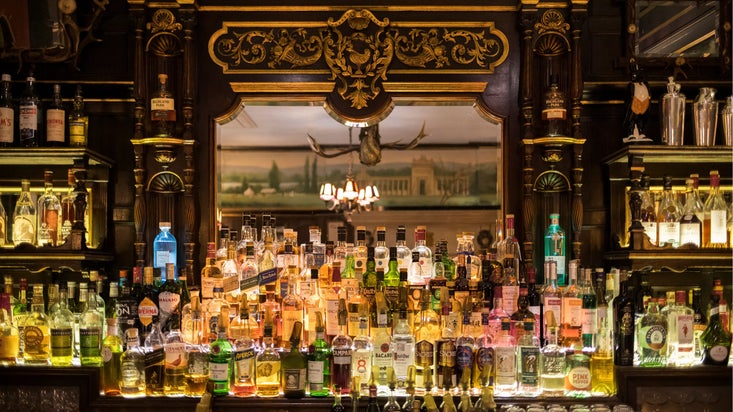 Traditional cocktail bar with ornate dark wood panelling, a large decorative mirror, and a wide selection of illuminated spirits and liqueurs displayed on shelves and behind the counter.